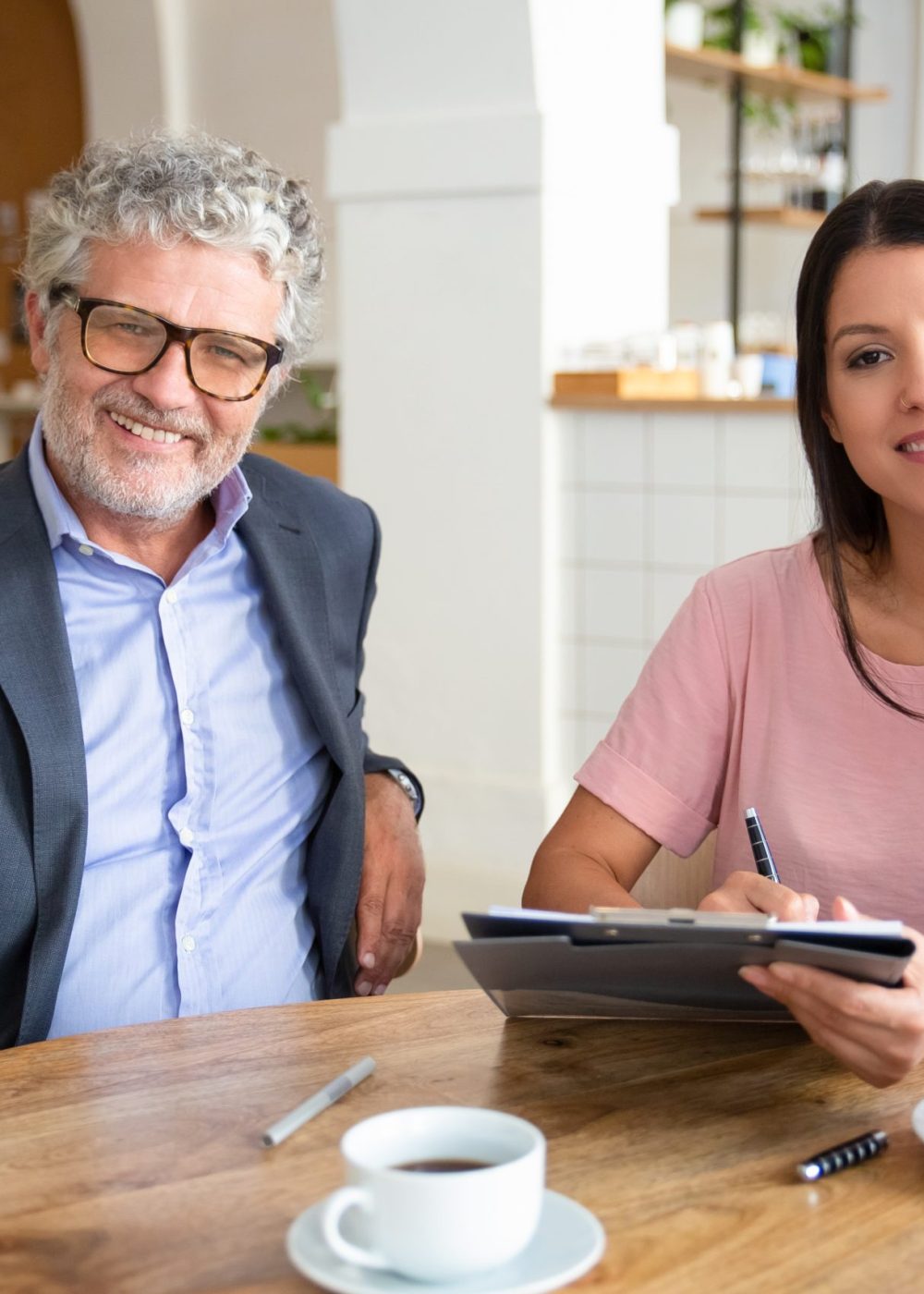 Agent and customer meeting over cup of coffee at co-working, sitting at table, holding documents, looking at camera and smiling. Medium shot, front view. Paperwork or cooperation concept