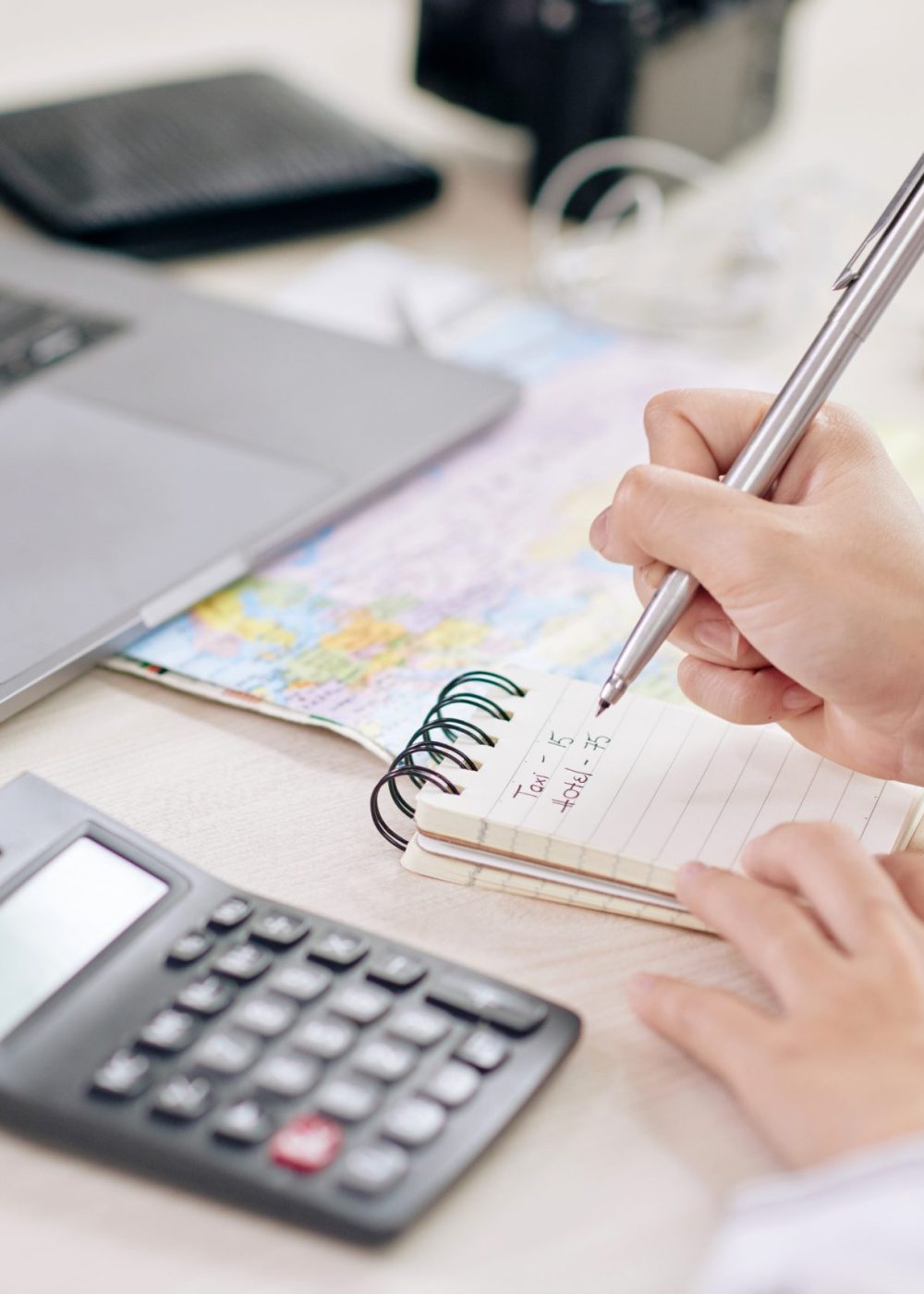 Crop shot of woman taking notes in notepad sitting at desk with laptop and calculating machine